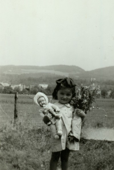 Girl with doll and bouquet outdoors: A young girl with dark hair and a large bow holding a doll in her left hand and a bouquet of flowers in her right, with hills and a fence in the background