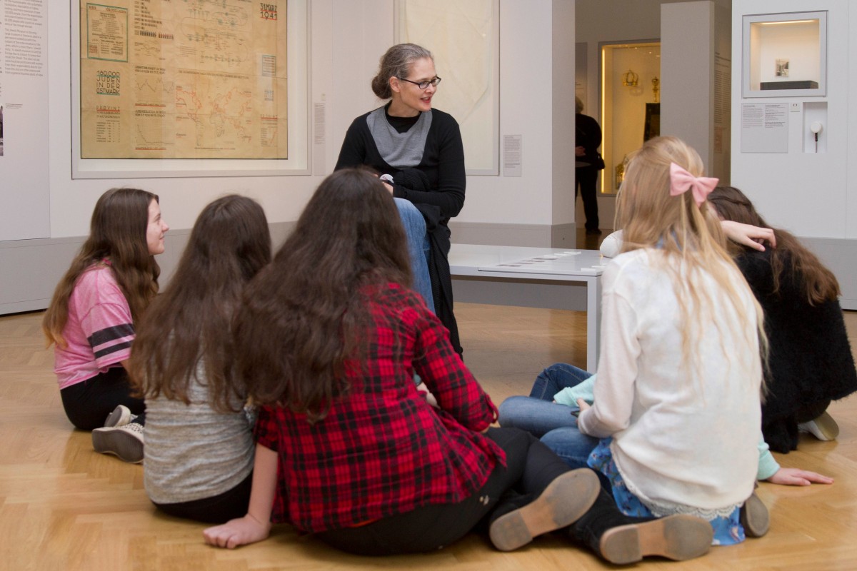 Museum tour with youths: Seven girls sitting on the floor of a museum room listening to a woman with glasses and black clothing sitting on a bench