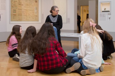 Führung im Museum mit Jugendlichen: Sieben Mädchen sitzen auf dem Boden eines Museumsraums, hören einer sitzenden Frau mit Brille und schwarzer Kleidung zu