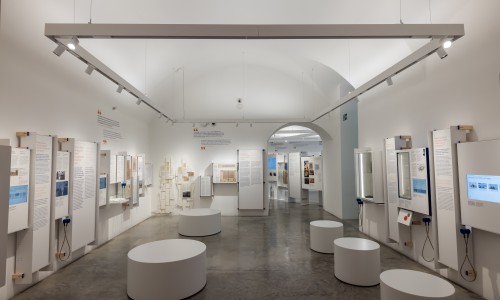 Langer Ausstellungsraum mit weißen Wänden und gebogenem Gewölbe, Informationspanelen an den Wänden und runden weißen Sitzgelegenheiten auf dem grauen Boden.
