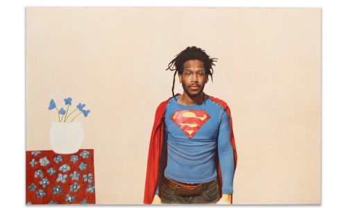 Mann mit Dreadlocks trägt Superman-T-Shirt und rote Umhang, steht vor beigem Hintergrund neben Tisch mit Vase und blauen Blumen.