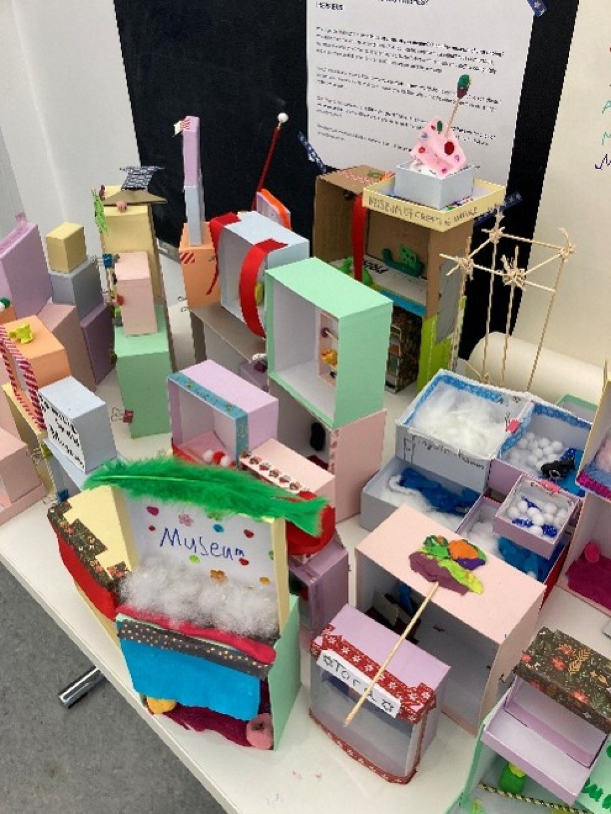 : Several colorful handmade models made of cardboard and paper are displayed on a table, including one labeled 'Museum'.