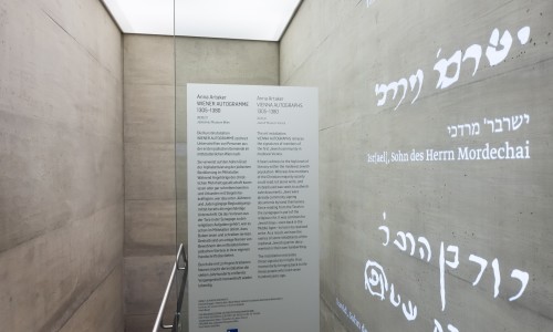 Treppenabgang mit Betonwänden, eine Glaswand mit Texttafel und projizierte hebräische und deutsche Schrift an der rechten Wand