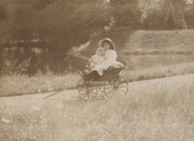 Children in antique baby carriage on country path: Two children sit in an antique four-wheeled baby carriage on a grassy path with trees in the background