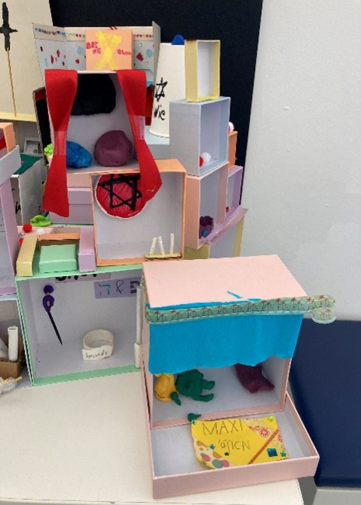 : Model of a multi-story dollhouse made of cardboard with colorful details, curtains, and small figures.