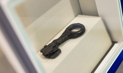 Ein alt aussehender, schwarzer Metall- oder Eisen-Schlüssel in einem weißen Ausstellungsrahmen.