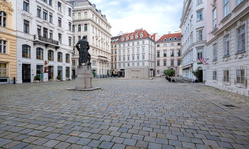 Großer gepflasterter Platz mit Statue auf weißem Sockel in der Mitte, umgeben von historischen mehrstöckigen Gebäuden mit weißen Fassaden.
