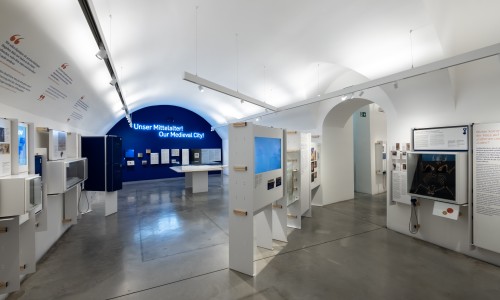 Ausstellungsraum im Jüdischen Museum Wien mit Textwänden und Exponaten zur mittelalterlichen Stadt, weiße Wände, blauer Hintergrund mit Text 'Unser Mittelalter! Our Medieval City!'