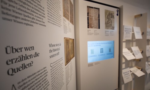 Ausstellungswand mit Texten und Bildern, darunter 'Über wen erzählen die Quellen?' und 'Whose story do the historical sources tell?'