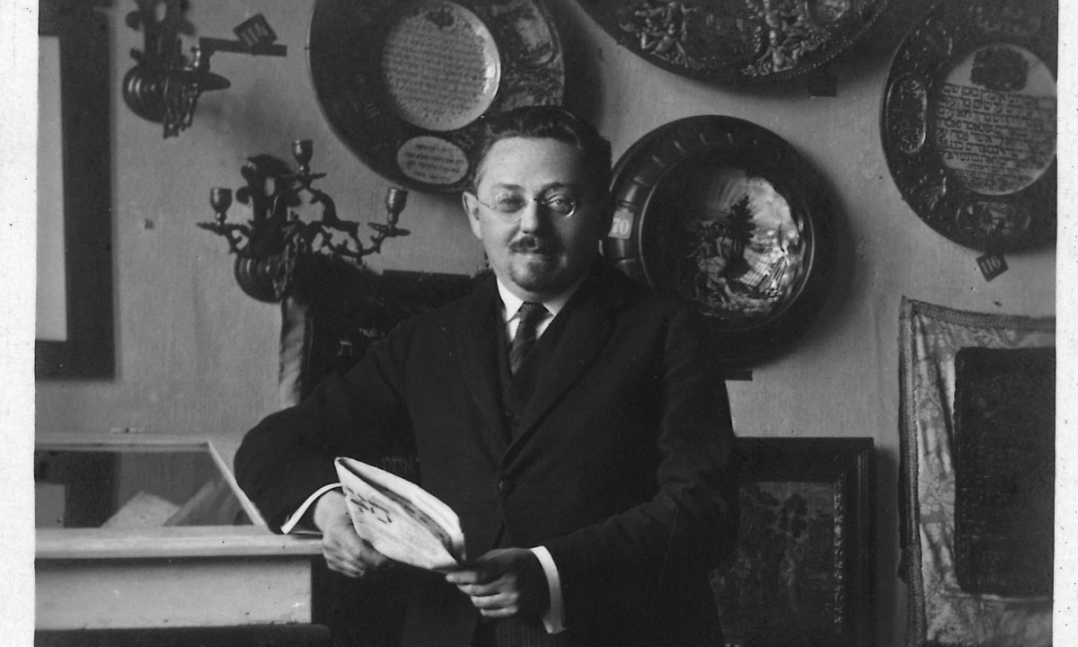 A man wearing glasses and dressed in a three-piece suit stands in an exhibition space, holding a newspaper in his hands.