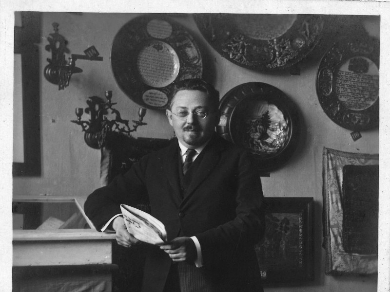 A man wearing glasses and dressed in a three-piece suit stands in an exhibition space, holding a newspaper in his hands.
