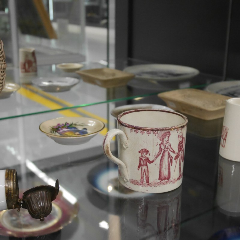 Vitrine mit bemalten historischen Keramik- und Porzellangegenständen, darunter ein Bierkrug, Tassen mit Figurenmotiven und dekorative Teller