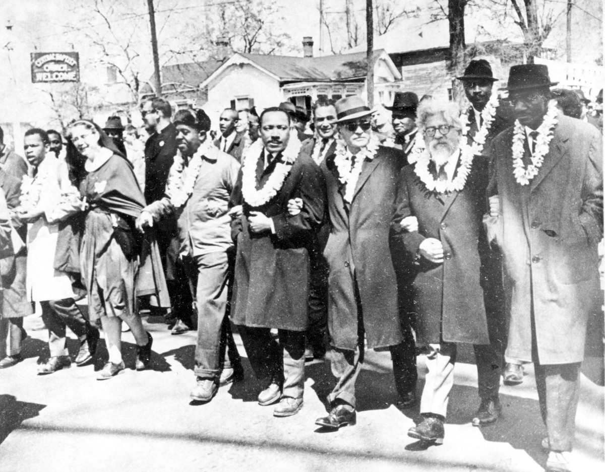 : Group of people wearing coats and flower garlands standing arm in arm on a street. Martin Luther King and Rabbi Heschel