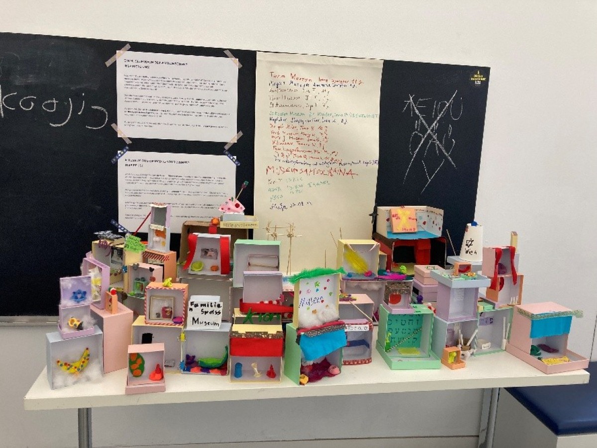 : Several small, colorful model houses made of cardboard and paper are displayed on a white table in front of a blackboard with handwritten texts and drawings.