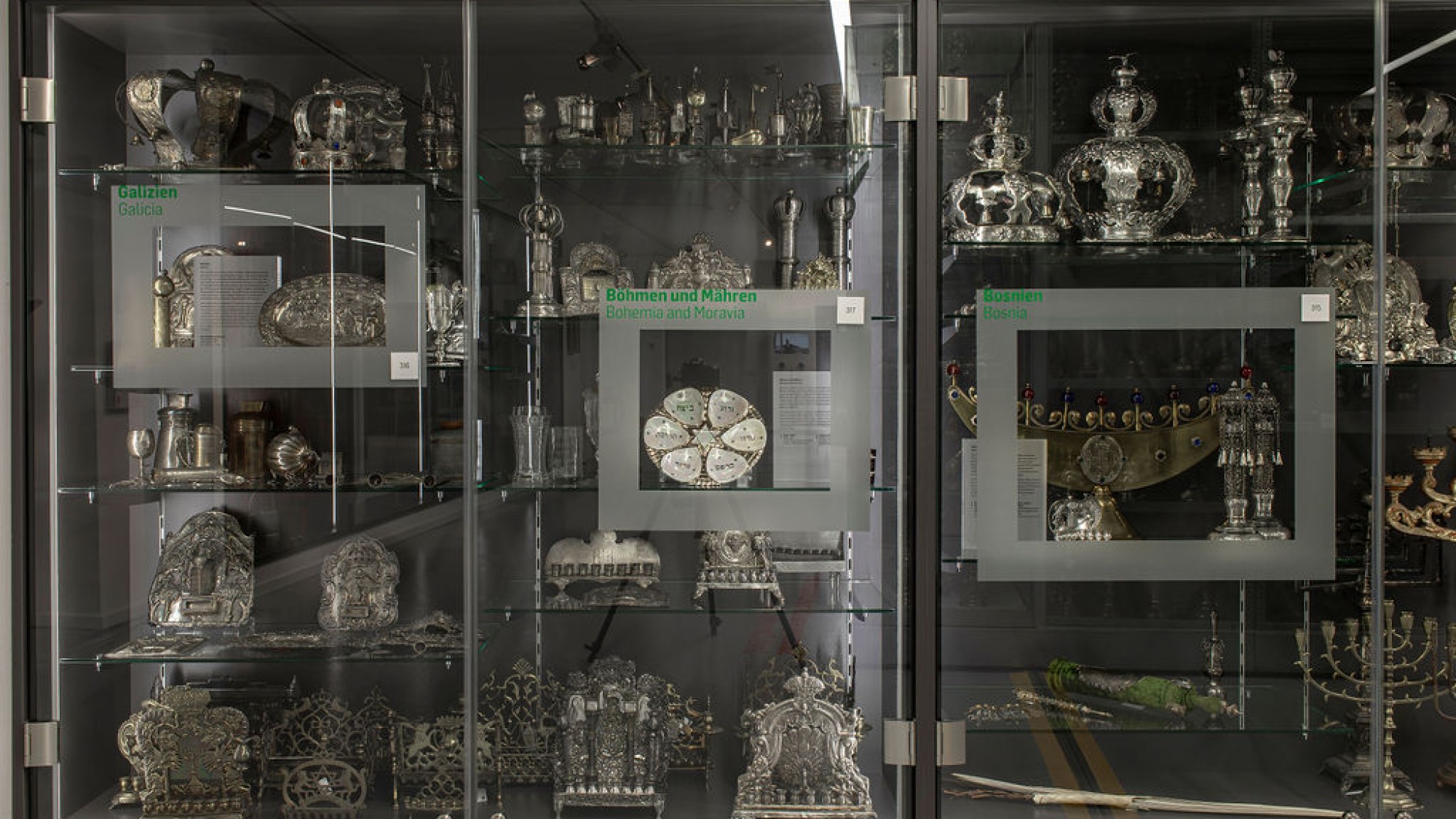 Display cases with Jewish ritual objects from various regions: Three glass cases containing silver Jewish ritual objects from Galicia, Bohemia and Moravia, and Bosnia, including crowns, candlesticks, and book holders.