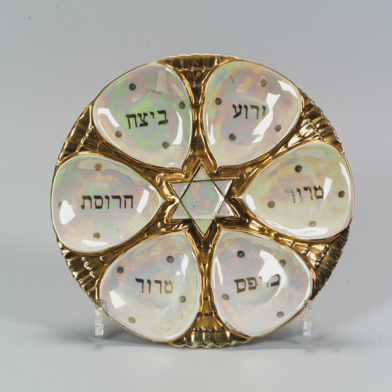 Goldener runder Seder-Teller mit sechs regenbogenfarben schimmernden, tropfenförmigen Vertiefungen, beschriftet mit hebräischen Namen traditioneller Speisen, in der Mitte ein Davidstern