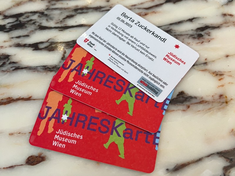 Jahreskarten und Namenskarte des Jüdischen Museums Wien: Zwei rote Jahreskarten des Jüdischen Museums Wien mit Schrift und bunten Silhouetten liegen auf einem marmorierten Untergrund. Obenauf liegt eine weiße Karte mit Text und dem Namen Berta Zuckerkandl.