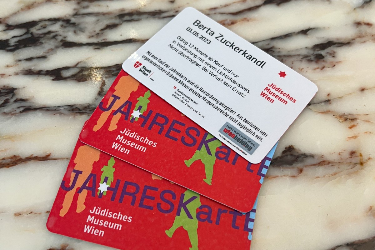 Annual passes and named card of the Jewish Museum Vienna: Two red annual passes for the Jewish Museum Vienna with text and colorful silhouettes lie on a marbled surface. On top is a white card with text and the name Berta Zuckerkandl.