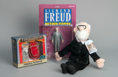 Sigmund Freud Toy Figures: Three Sigmund Freud figures: an action figure in packaging, a finger puppet set with Freud and a red chair, and a seated plush figure wearing a black suit and glasses.