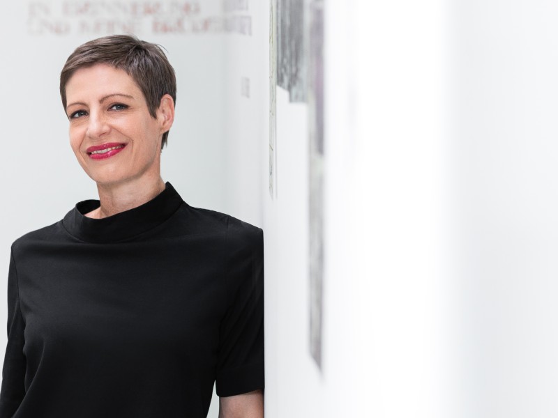 Frau mit kurzem braunem Haar und rotem Lippenstift lehnt an weißer Wand in Galerie mit sichtbaren Kunstwerken