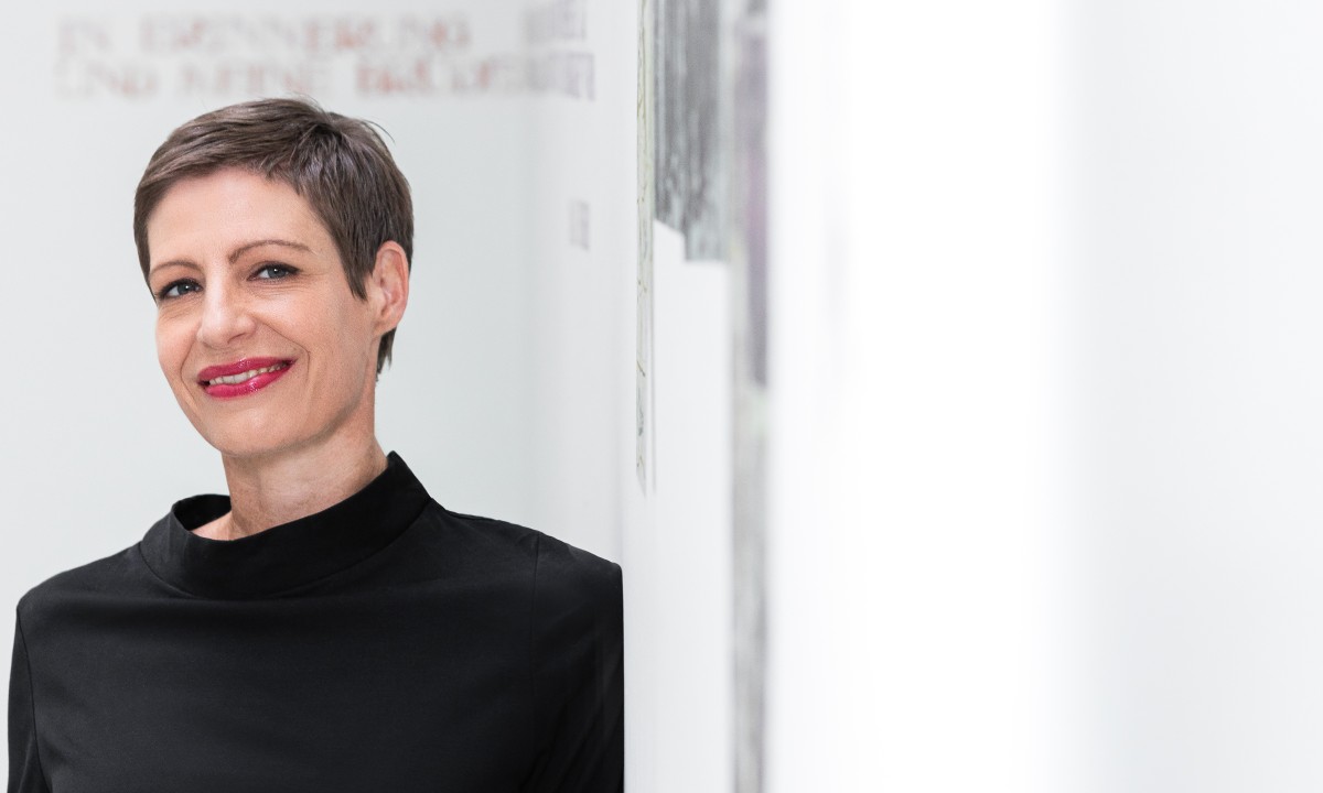 Woman with short brown hair and red lipstick leaning against white wall in gallery with visible artworks