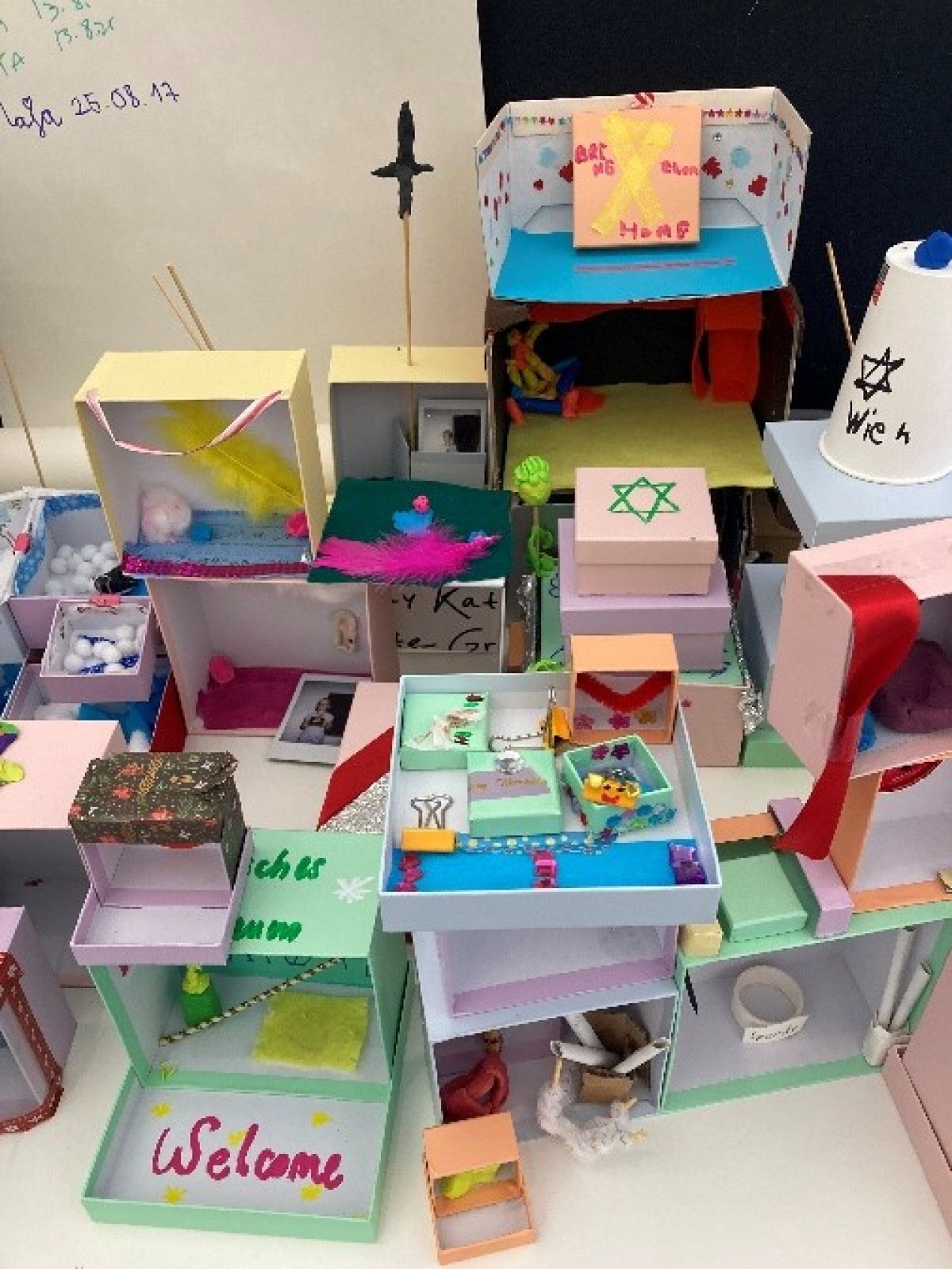 : Several colorful handmade miniature models made of paper and cardboard, including one with a green Star of David and one with the word 'Welcome'.