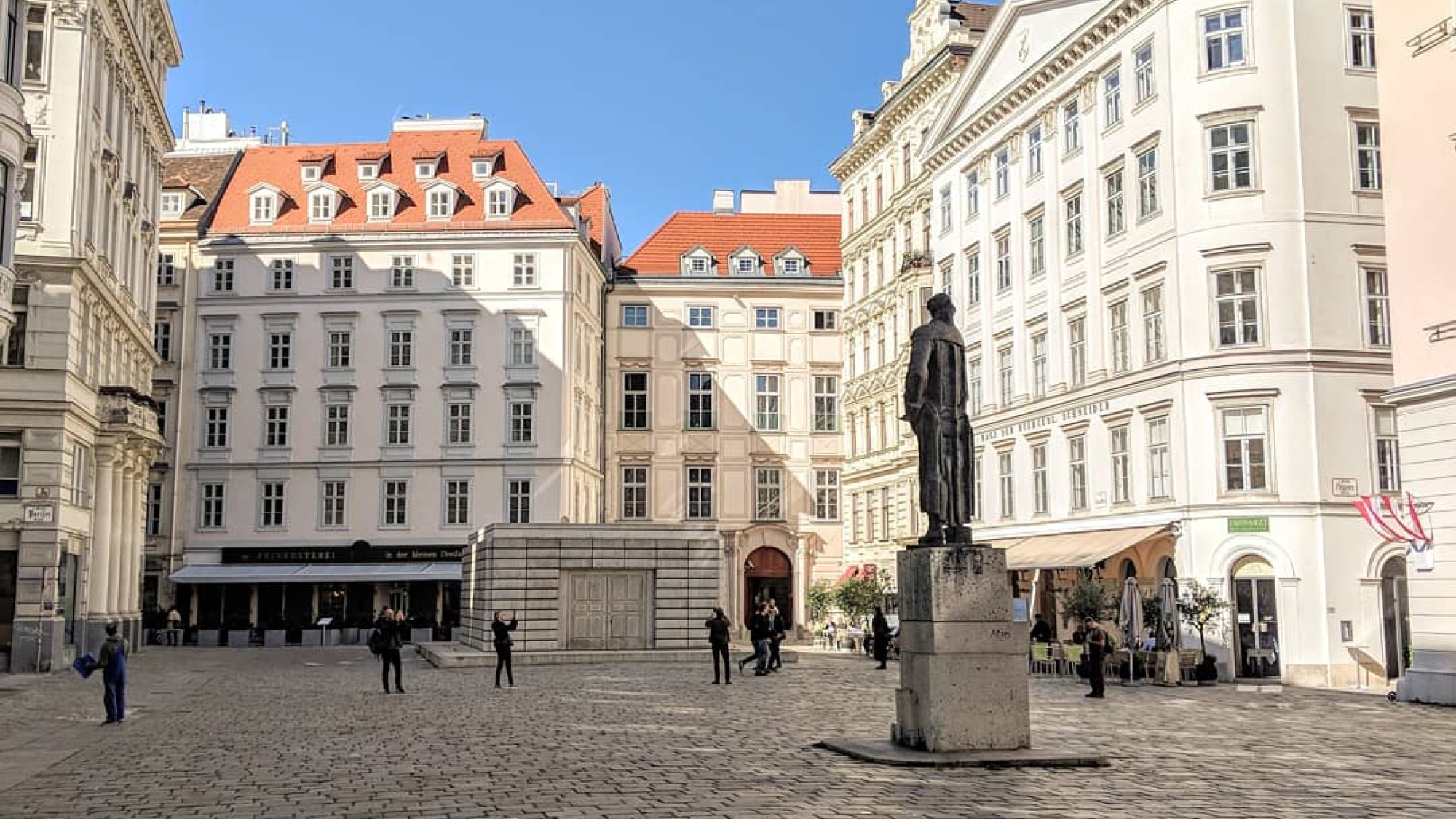 Pestsäule auf einem historischen Platz in Wien: Pestsäule mit Statue in einem gepflasterten Platz umgeben von historischen weißen Gebäuden mit roten Dächern bei klarem blauem Himmel