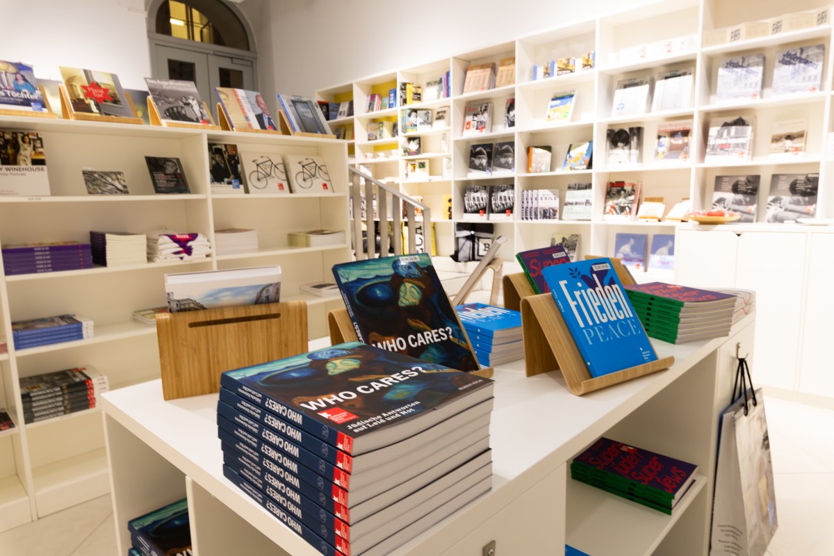 Museumsshop mit Büchern: Innenansicht des Museumsshops mit Regalen voll Bücher und Zeitschriften, im Vordergrund Stapel von Büchern mit dem Titel 'WHO CARES?' und 'Frieden PEACE'