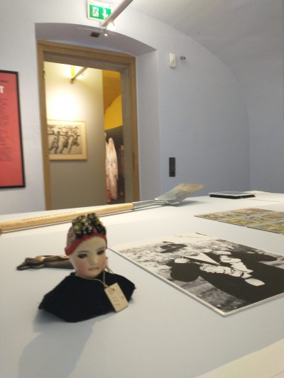 Exhibition objects at the Jewish Museum Vienna: Exhibition with a small doll bust wearing red headgear and black clothing, an old black-and-white photograph, and other objects on a white table in a bright room.