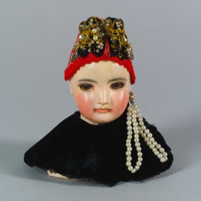 Doll head with red cap and pearls: Head of a doll figure with rosy cheeks, black cloak, and red cap decorated with pearls