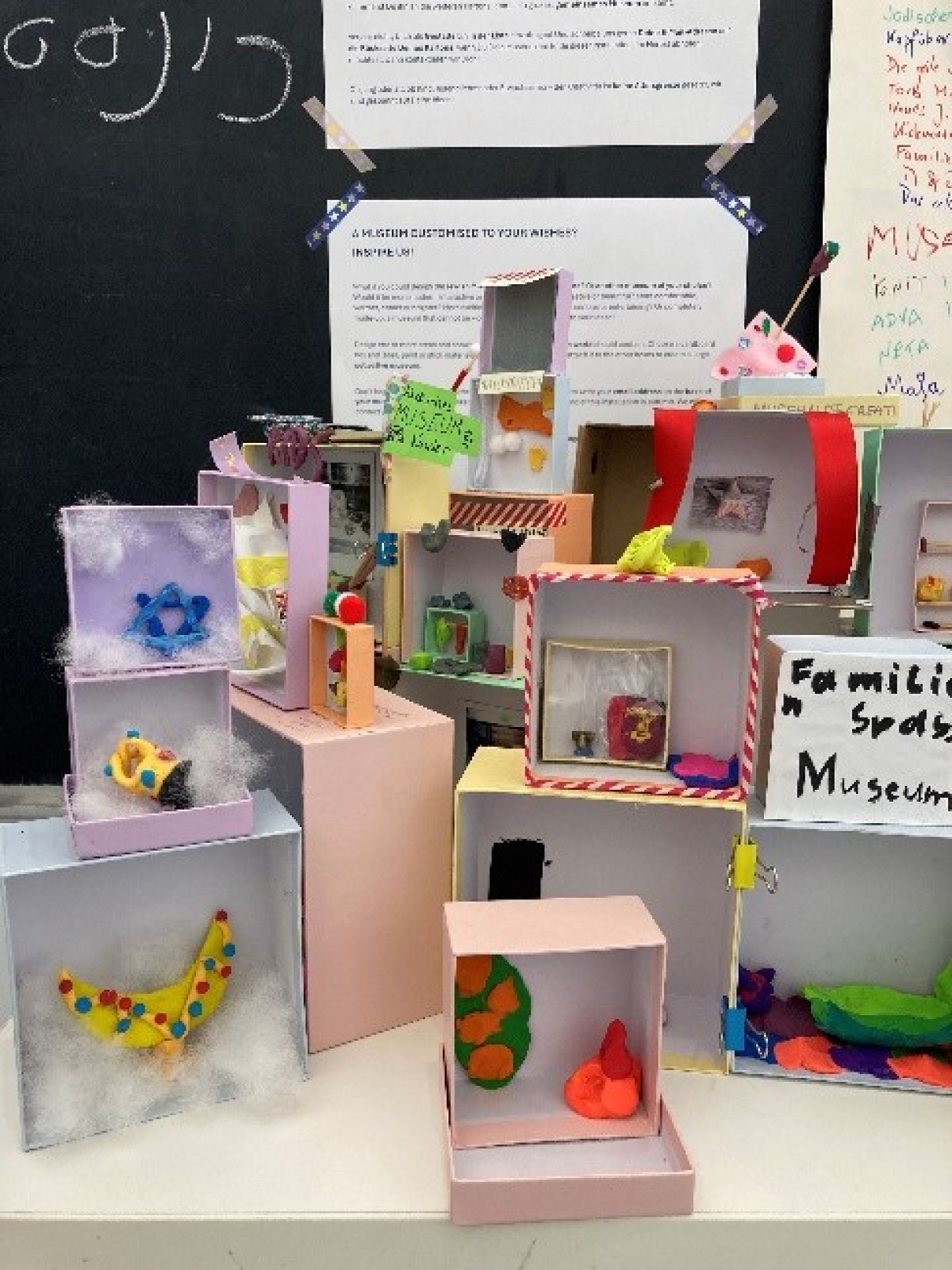 : Several small colorful dioramas made from cardboard boxes with various figures and decorations, displayed on a table in front of a blackboard and white text sheets.