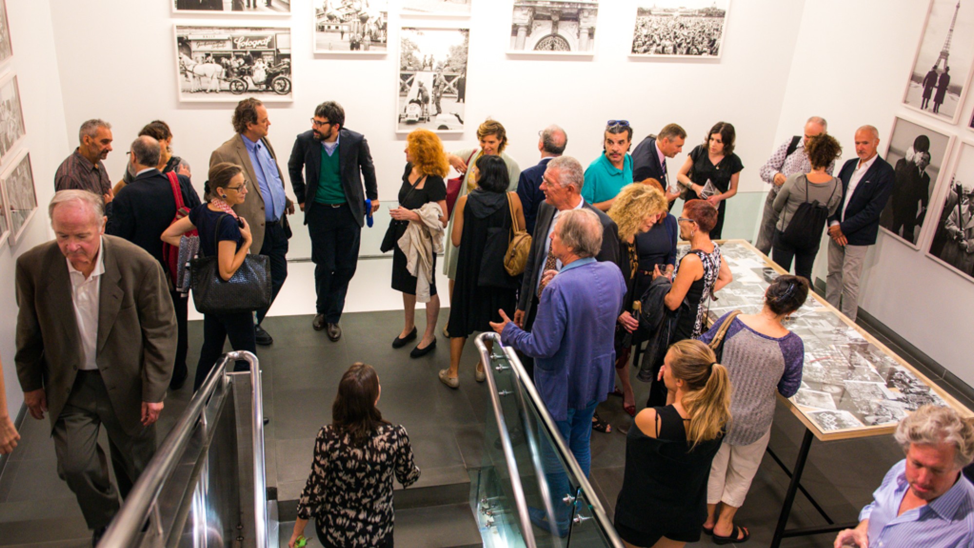 Museum visitor group: A group of people in a museum gathered around black and white photographs on walls and a long table in the room, engaging in conversation.