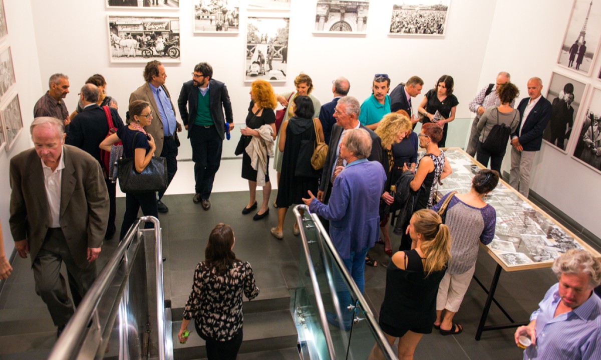 Besuchergruppe im Museum: Eine Gruppe von Menschen in einem Museum, die sich vor schwarzen und weißen Fotografien an den Wänden und auf einem langen Tisch im Raum unterhalten.