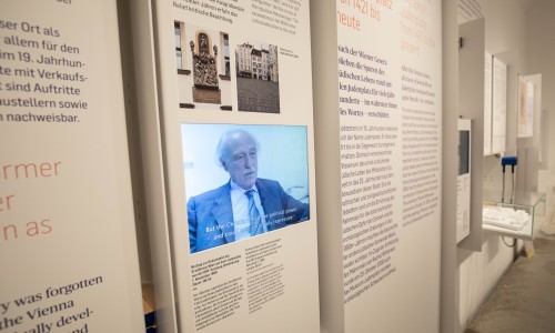 Museumsausstellung mit Texttafeln und einem Bildschirm, der ein älteren Mann in einem Anzug zeigt, der spricht und Untertitel auf Englisch trägt.