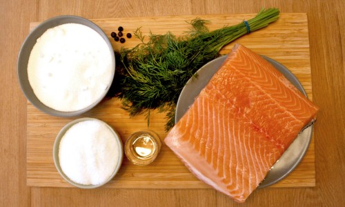 : A wooden board with a piece of salmon fillet on a plate, two bowls containing sugar and salt, a small glass with liquid, a bunch of dill, and some juniper berries.