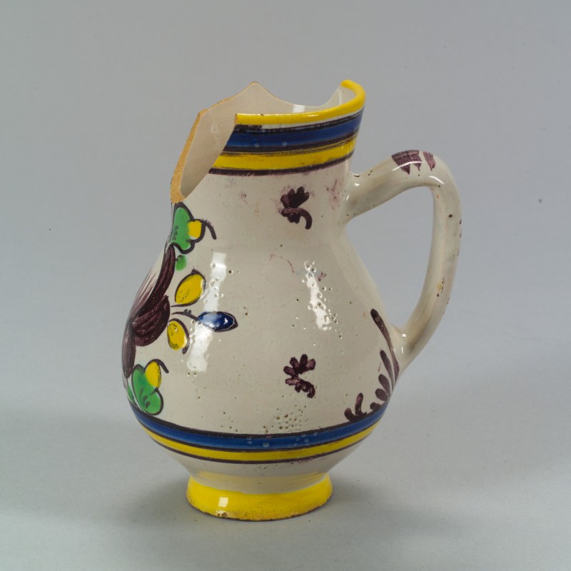 Beschädigte weiße Keramikkanne mit Henkel, gelbem Rand und handgemalten blauen, gelben und grünen Blumenmotiven