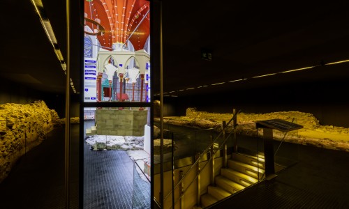 Unterirdischer Ausstellungsraum mit archäologischen Mauerruinen, beleuchteter Treppe und digitaler Infostele mit Bild einer historischen Bauwerksrekonstruktion