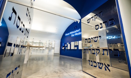 Blick in den Ausstellungsraum der Mittelalter-Ausstellung mit blauer Wand, Text 'Unser Mittelalter! Our Medie' und spiegelnden Tafeln mit hebräischen Buchstaben