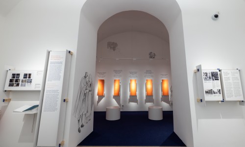 Ausstellungsraum mit weißem Bogen, fünf vertikal-orangen beleuchteten Informationstafeln an der Rückwand, zwei weißen Sitzhockern und Wandgrafik mit zwei historischen Figuren