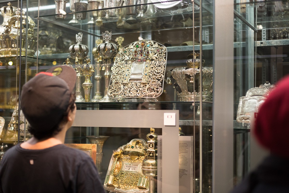 Vitrine mit jüdischen Ritualobjekten: Zwei Personen betrachten eine Vitrine mit verzierten silbernen jüdischen Ritualobjekten, darunter Kronen und Torarollenhüllen.