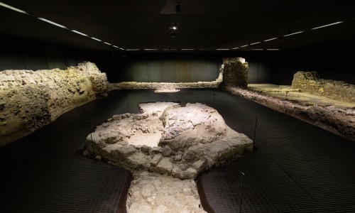Archäologische Überreste von Steinmauern und Fundamenten in einem dunklen Ausstellungsraum mit gezielter Beleuchtung