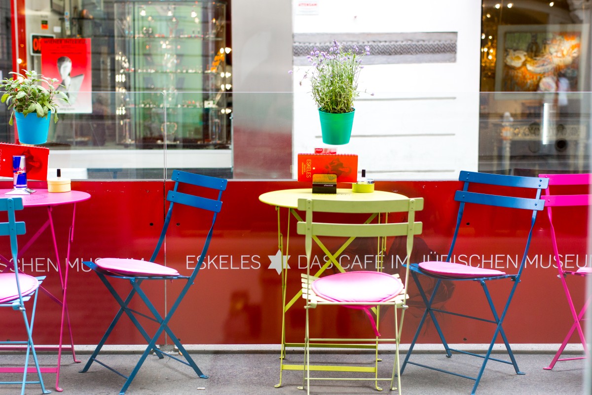 Outdoor seating at Eskeles Café: Colorful metal chairs and tables with light cushions in front of a glass wall reading 'ESKELES DAS CAFÉ IM JÜDISCHEN MUSEUM'