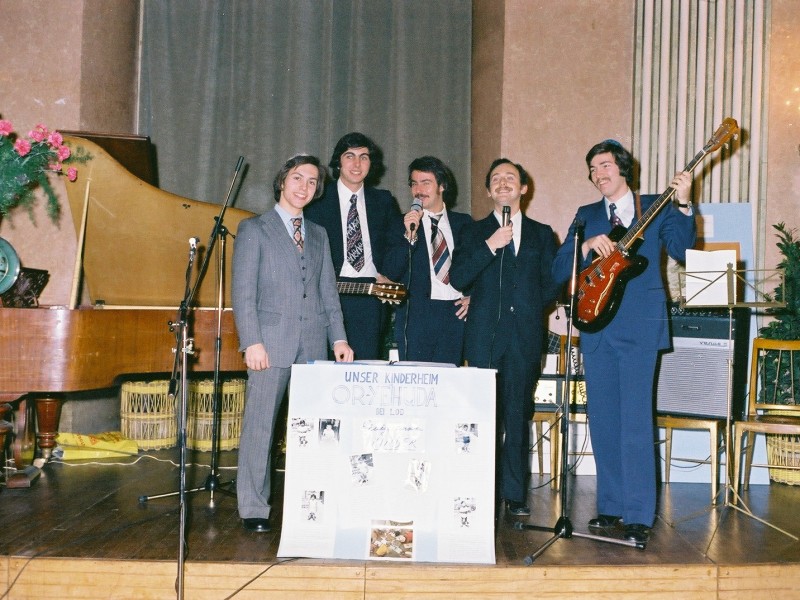 Fünf junge Männer in Anzügen auf einer Bühne mit Mikrofonen, Gitarre und Poster mit der Aufschrift ‚Unser Kinderheim OR-ZI-IDA‘