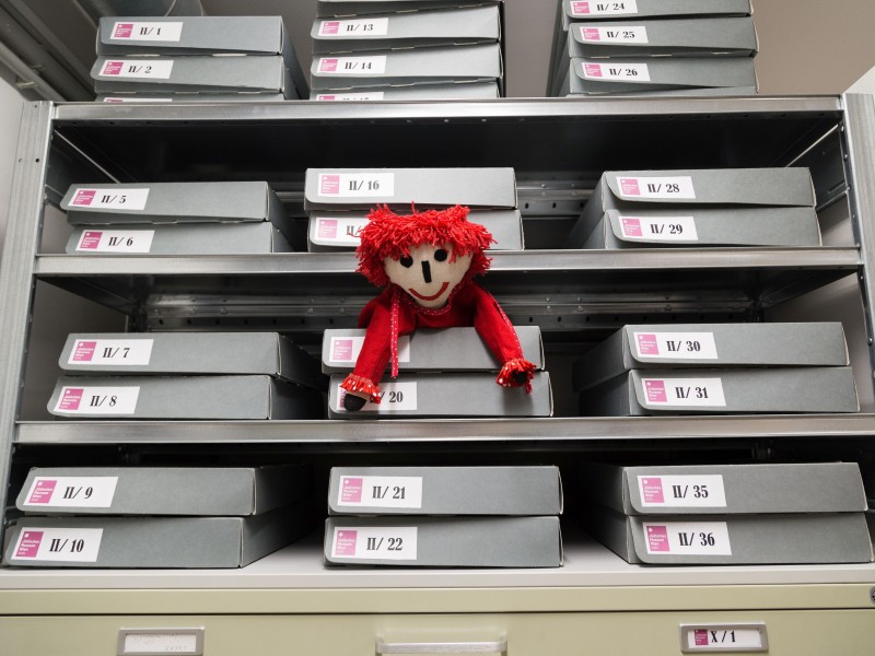 Archive shelf with doll: Shelf with gray archive boxes numbered II/1 to II/36 and X/1, with a red doll with red hair placed between boxes
