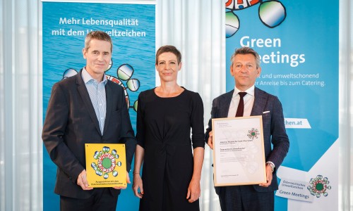 Drei Menschen stehen vor zwei Bannern, die Umweltthemen zeigen. Der Mann links hält ein gelbes Schild mit dem Österreichischen Umweltzeichen, der Mann rechts eine Urkunde.