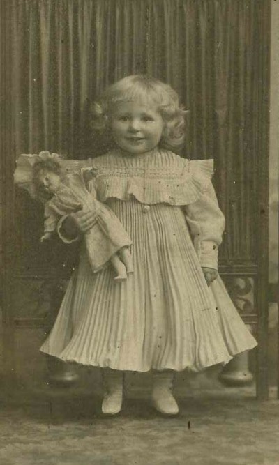 Child with doll in dress: Small child with curly hair wearing a long-sleeved, pleated dress with ruffles and buttons, holding a doll in the left hand