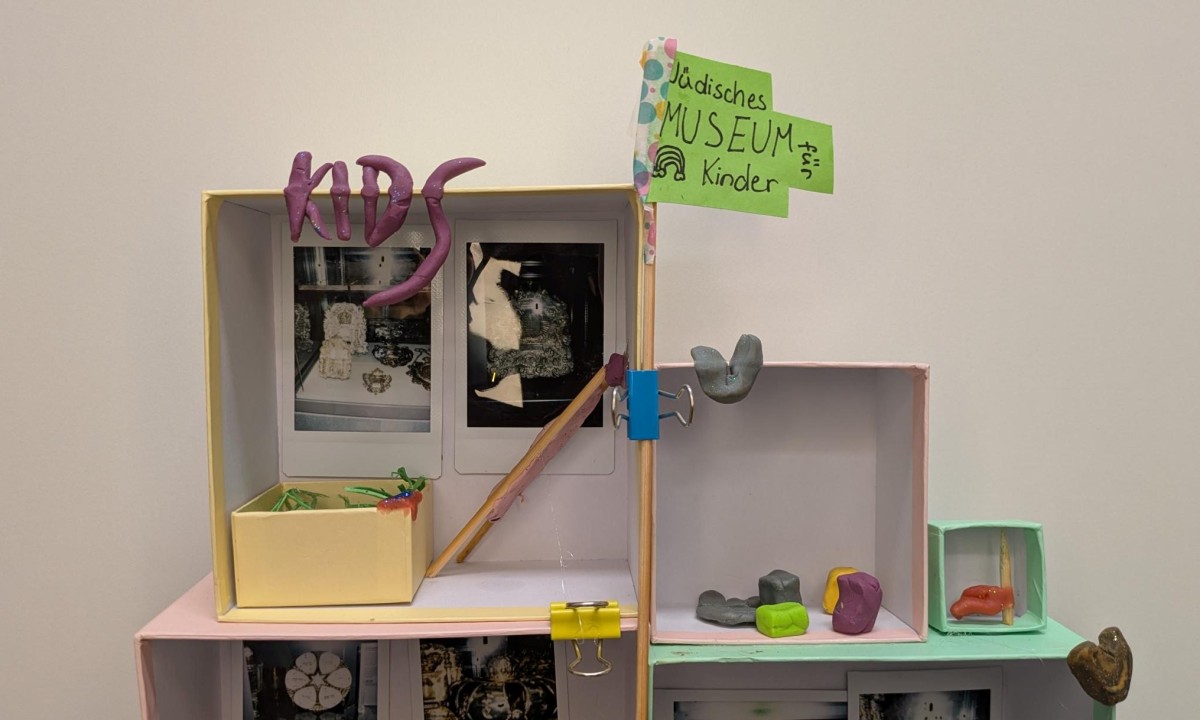 Small diorama with the word "KIDS" made of purple modeling clay, two photos in the background, and a small yellow box with green decoration in the foreground.