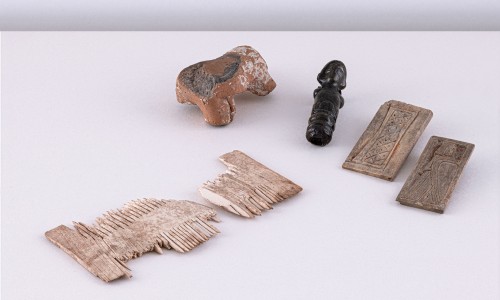 Drei antike Artefakte auf weißer Oberfläche: zerbrochener Elfenbeinkamm, braune Tonfigur in Tierform, kleine schwarze geschnitzte Figur, zwei braune rechteckige Reliefplatten mit Mustern