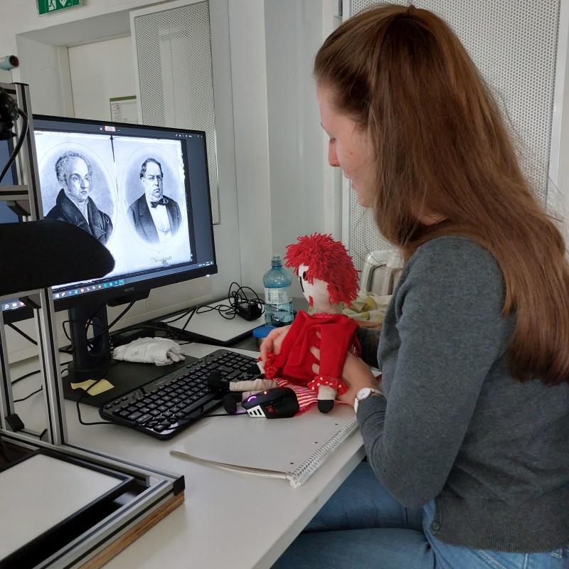 Frau am Computer mit historischer Darstellung und Puppe: Frau sitzt an einem Schreibtisch vor einem Computer mit zwei historischen Porträts auf dem Bildschirm, hält eine rote Puppe mit roten Haaren