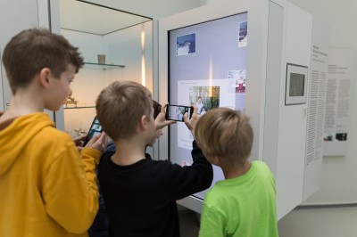 Kinder interagieren mit digitaler Museumsinstallation: Drei Jungen fotografieren mit Smartphones eine digitale Museumsinstallation mit Bildern und Erklärtexten